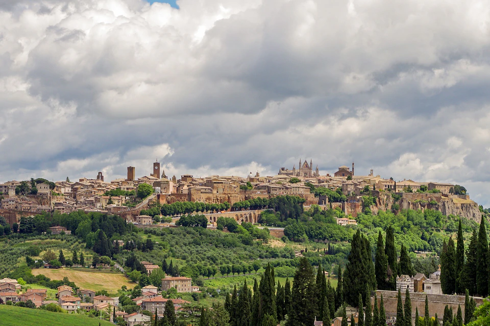 A medieval town with stone buildings and lush greenery, set against a backdrop of a cloudy sky.