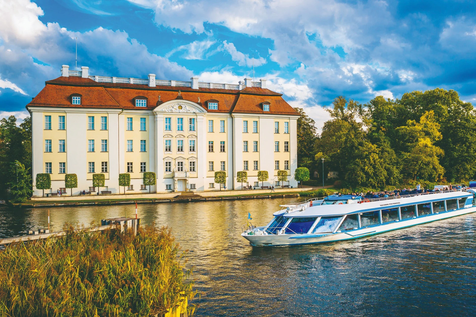 A river with a boat passing in front of a large, yellow, classical-style building under a partly cloudy sky.