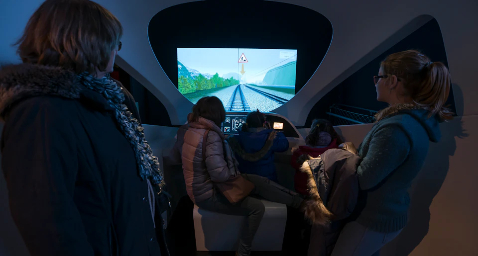 Three people seated in a dark exhibit, interacting with a railway simulator screen displaying train tracks and a warning sign.
