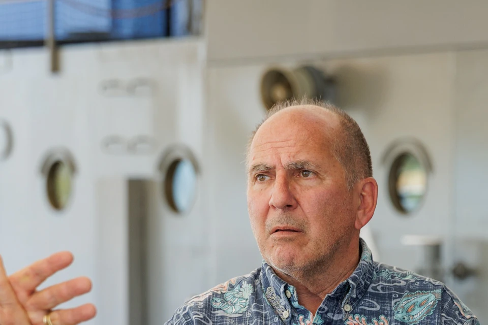 A man in a patterned shirt stands on a ship's deck, surrounded by others listening to a discussion.