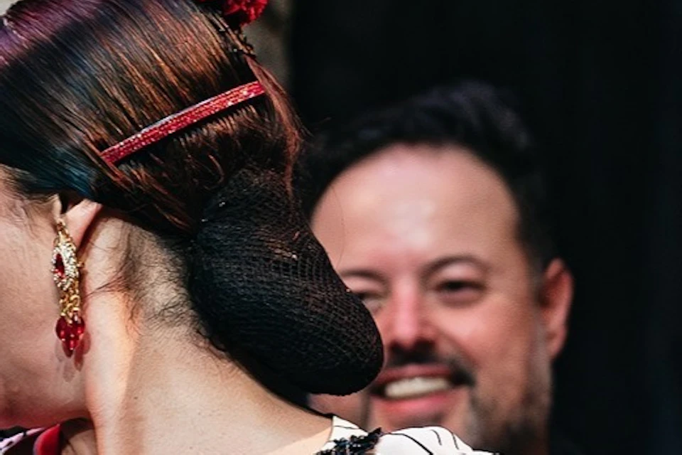 A flamenco dancer in a white dress performs on stage with musicians in the background, one clapping along.