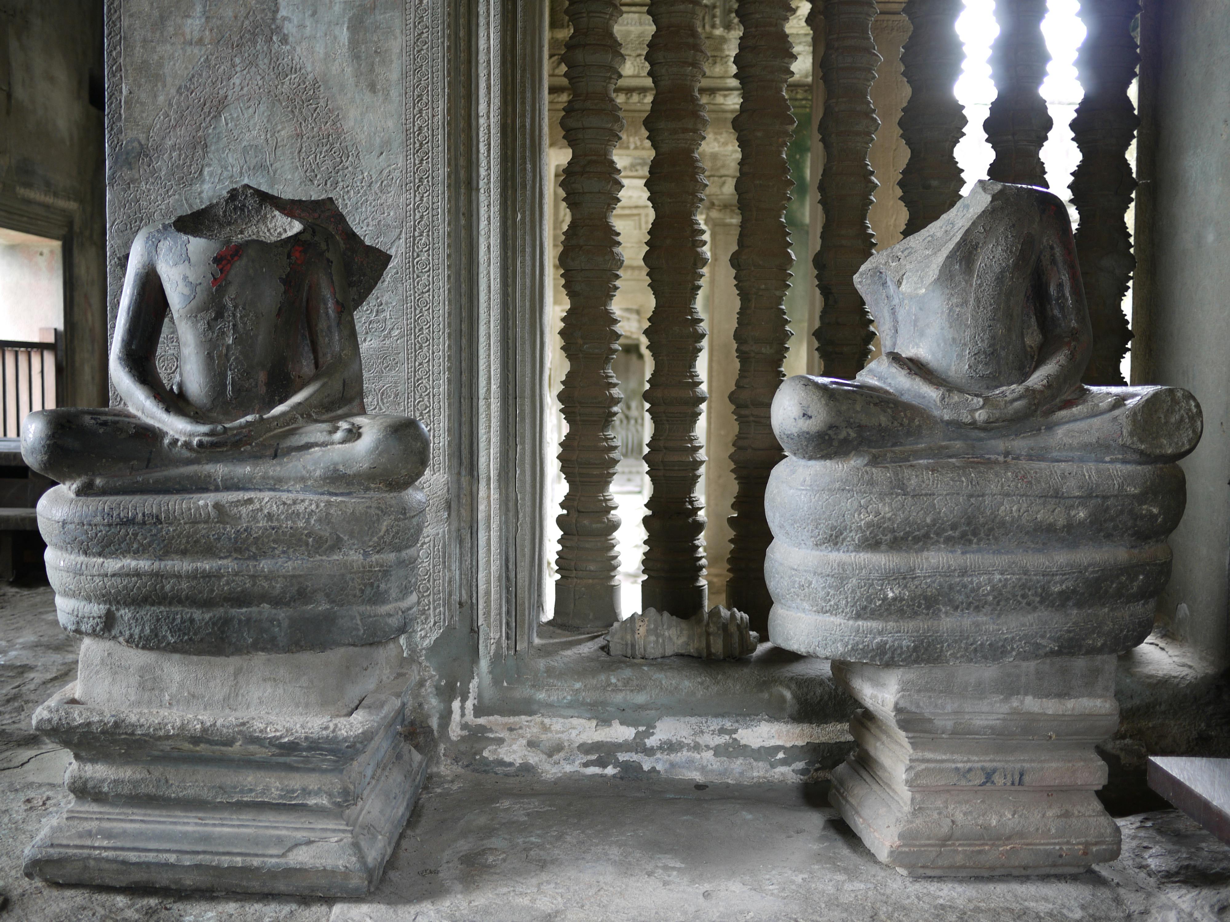 Two headless stone Buddha statues in seated positions, set against an ornate backdrop with intricate columns and weathered surfaces.