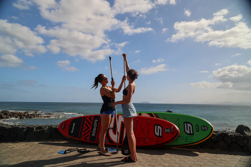 Two women standing on a paved path with paddleboards and paddles, adjusting paddle lengths near the ocean under a partly cloudy sky.