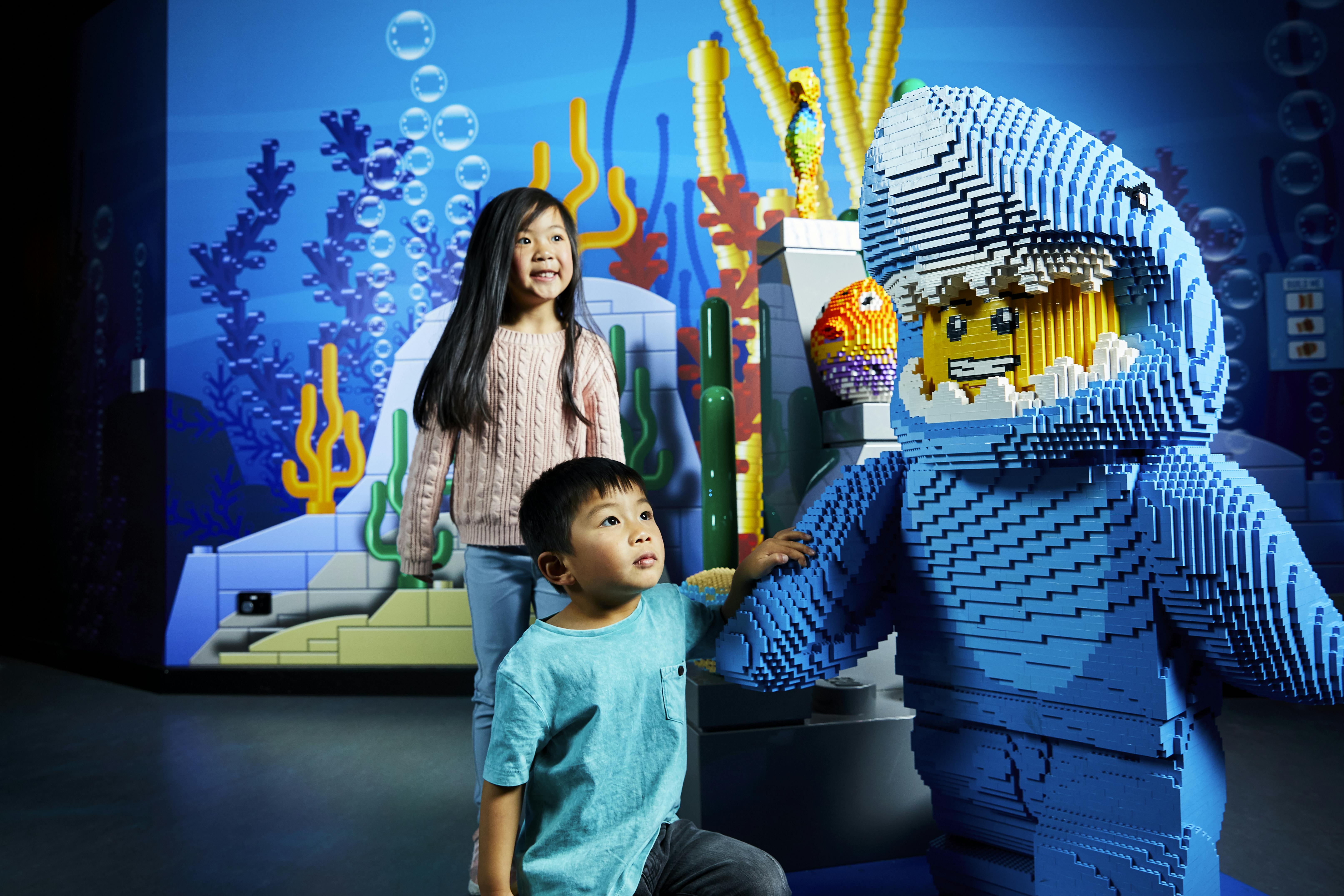 Two children smile in front of an underwater-themed LEGO display with colorful corals and a diver figure.