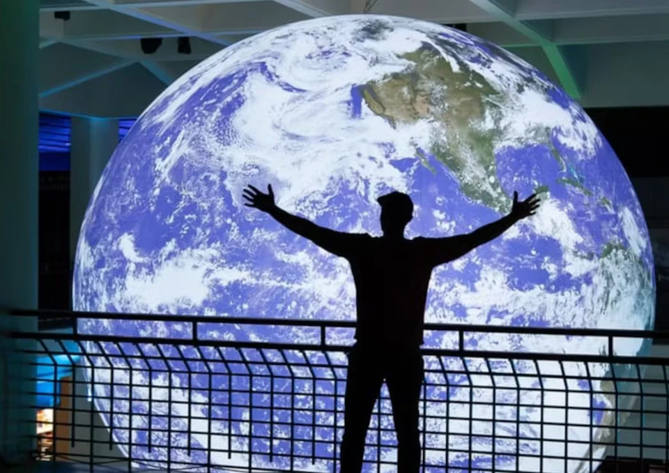 Silhouette of a person with arms outstretched in front of a large display of Earth in a modern exhibition space.
