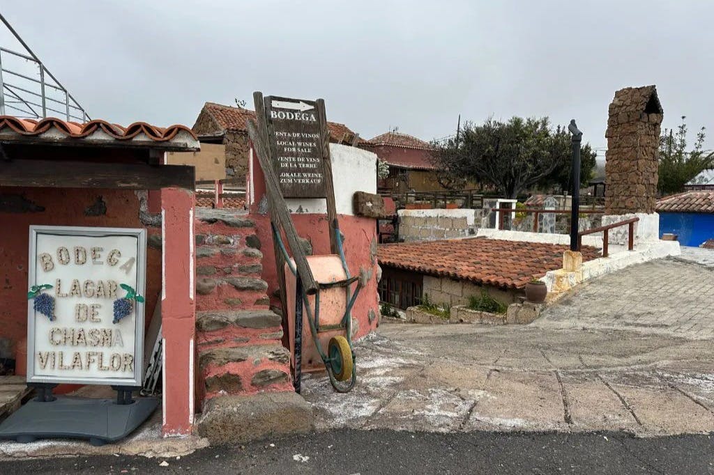 Rustikale Szene mit Backsteinmauern, einer alten Schubkarre, Steinstufen und einem Schild für eine Bodega. Ziegeldächer und bewölkter Himmel im Hintergrund.