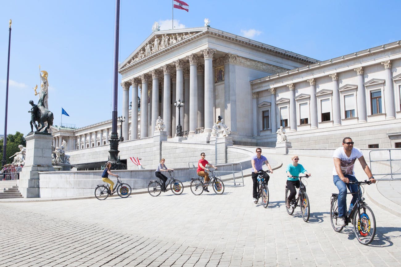 Lidé jezdící na kole před velkou budovou v klasickém stylu se sloupy a sochami. Ze střechy budovy vlaje vlajka.
