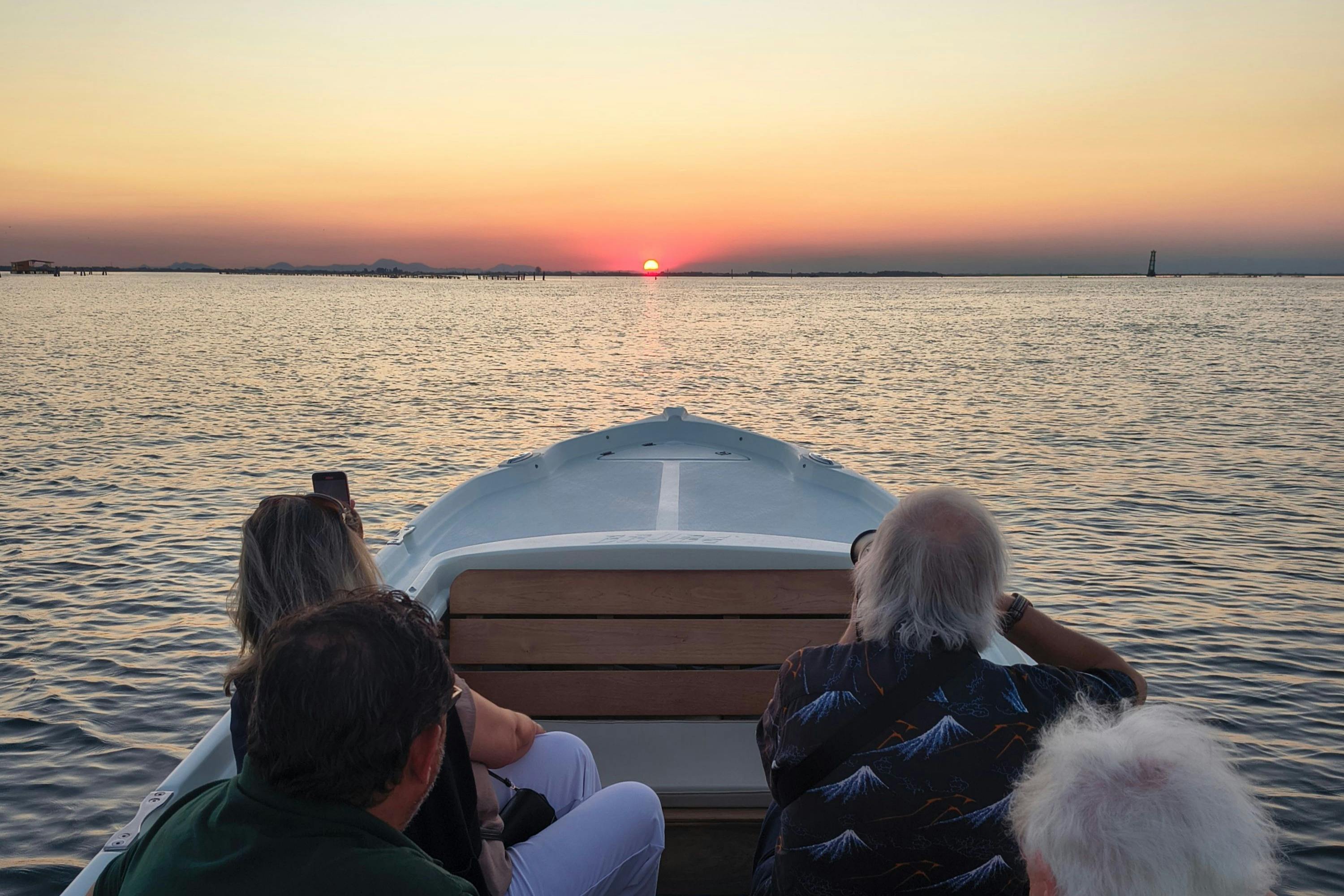 Passagiers op een boot kijken naar een zonsondergang boven kalm water, met de zon laag aan de horizon en een heldere hemel erboven.