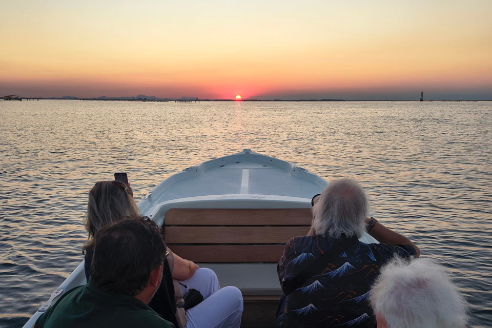 Passengers on a boat watch a sunset over calm water, with the sun low on the horizon and a clear sky above.