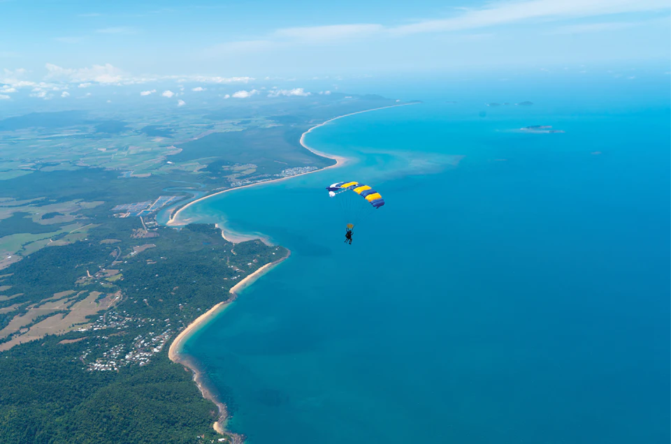 A person parachuting over a coastal landscape with lush greenery and blue ocean waters under a clear sky.