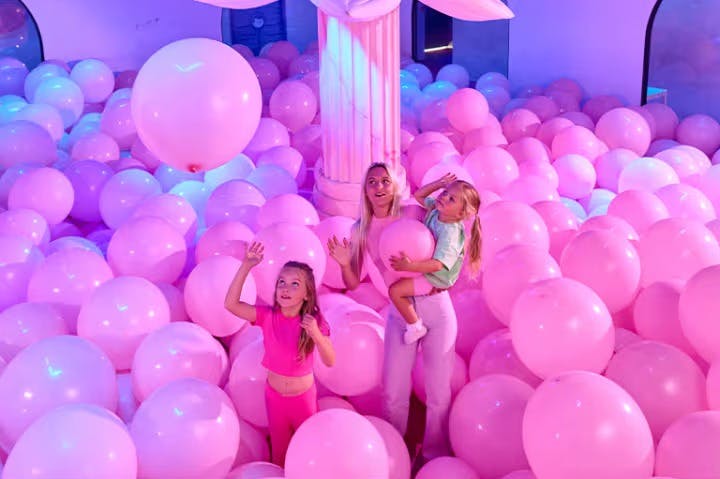 Three girls, surrounded by large pink balloons, play in a room with a white column and soft blue and purple lighting.