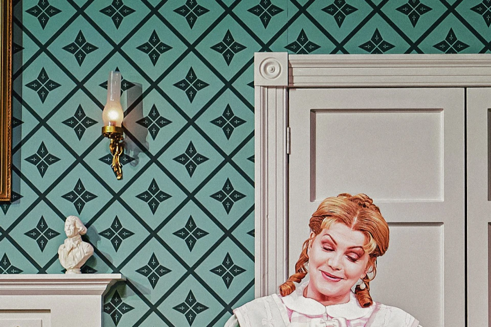 Two people in period attire sit in a historical room with patterned walls, a fireplace, and a portrait of George Washington.