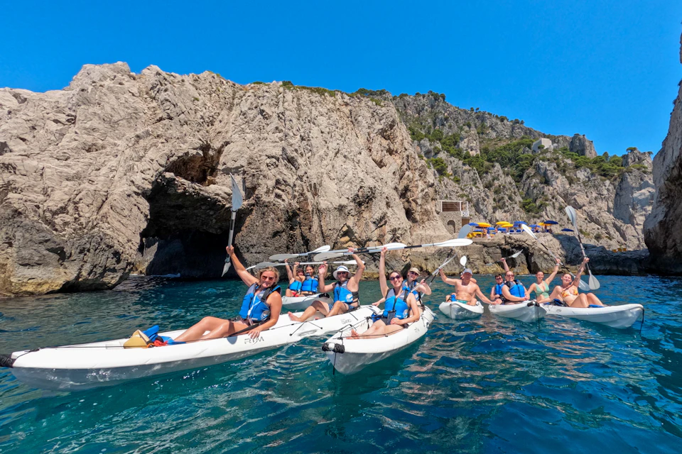 A group of people wearing life jackets paddle kayaks on clear blue water near rocky cliffs under a bright blue sky.
