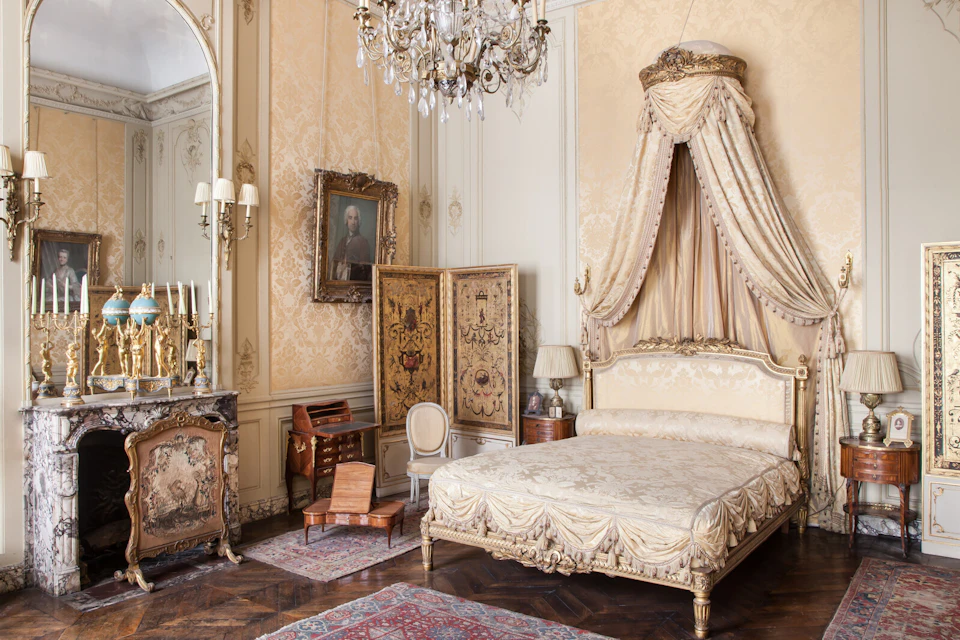 Ornate bedroom with a chandelier, canopy bed, antique furniture, large mirror, carpet, and a portrait on the wall.