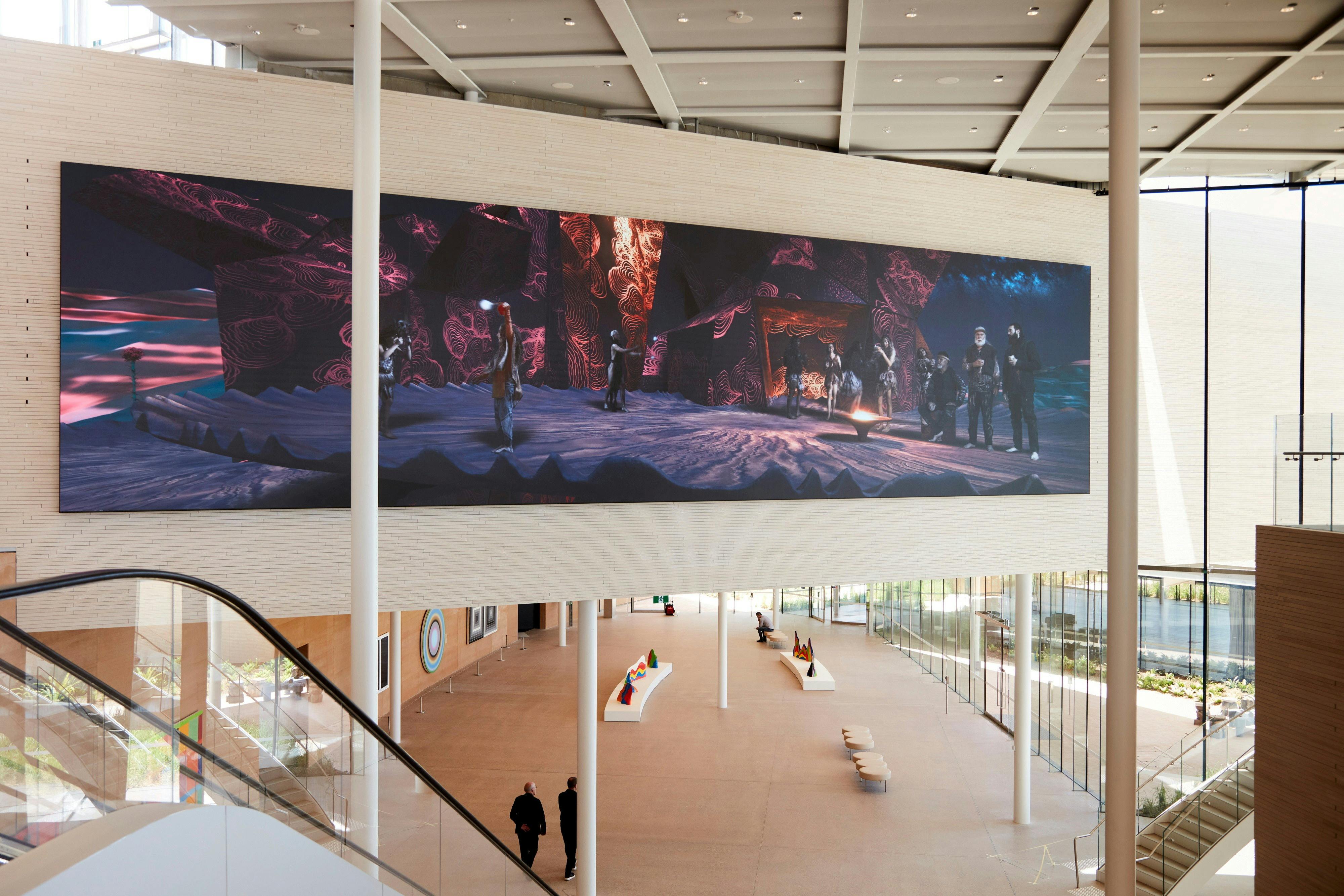 Spacious modern atrium with a large digital mural on the wall, depicting people in a dramatic scene. Two people walk below.