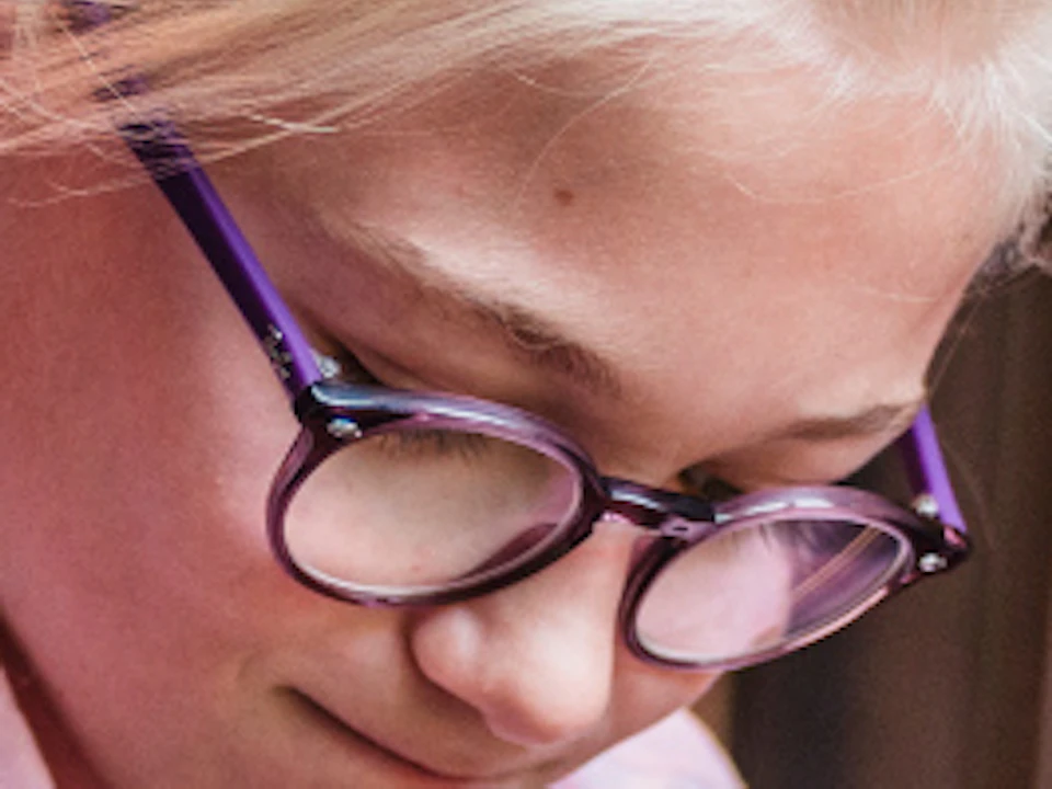 A girl wearing glasses and a pink shirt examines a metallic object on a table in a science exhibition setting.