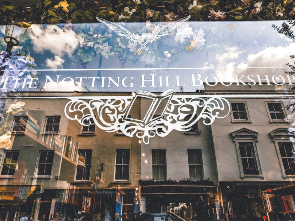 Storefront window of Notting Hill Books with decorative book engraving and street reflection of buildings and sky.