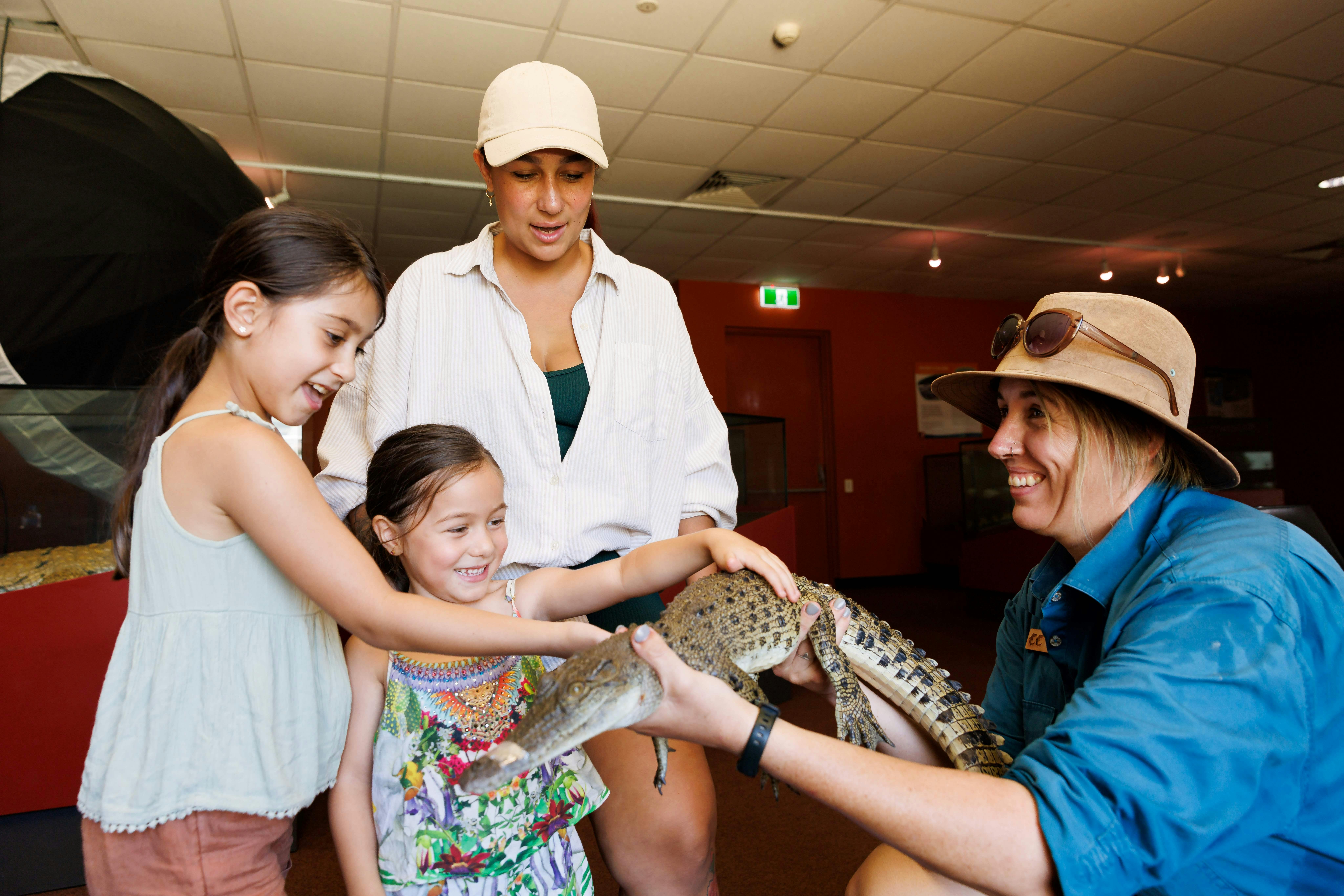 두 아이와 한 여성이 실내에서 동물원이 들고 있는 작은 악어를 쓰다듬고 있습니다.