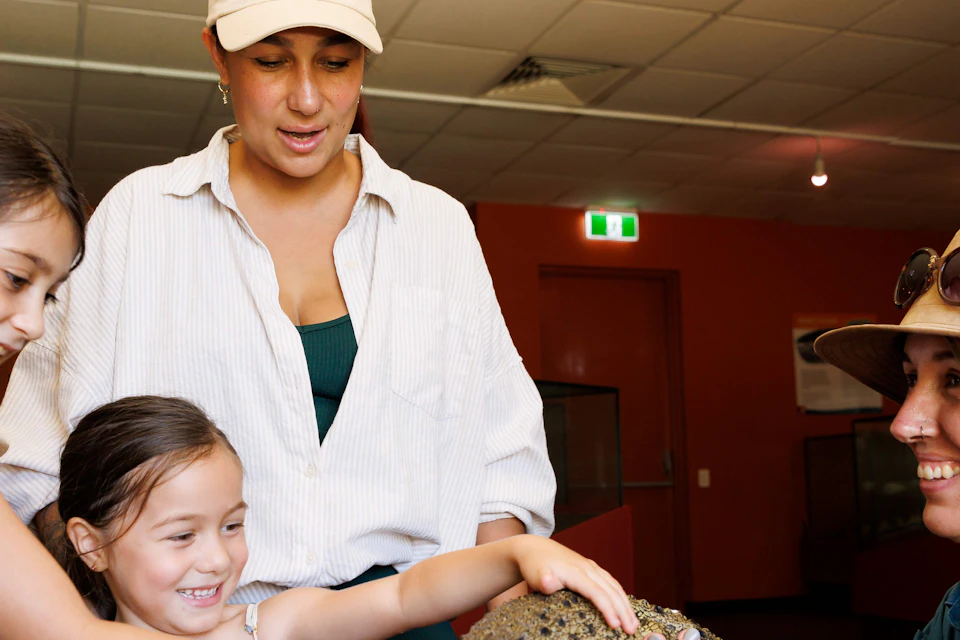 Two children and a woman pet a small crocodile held by a zookeeper indoors.