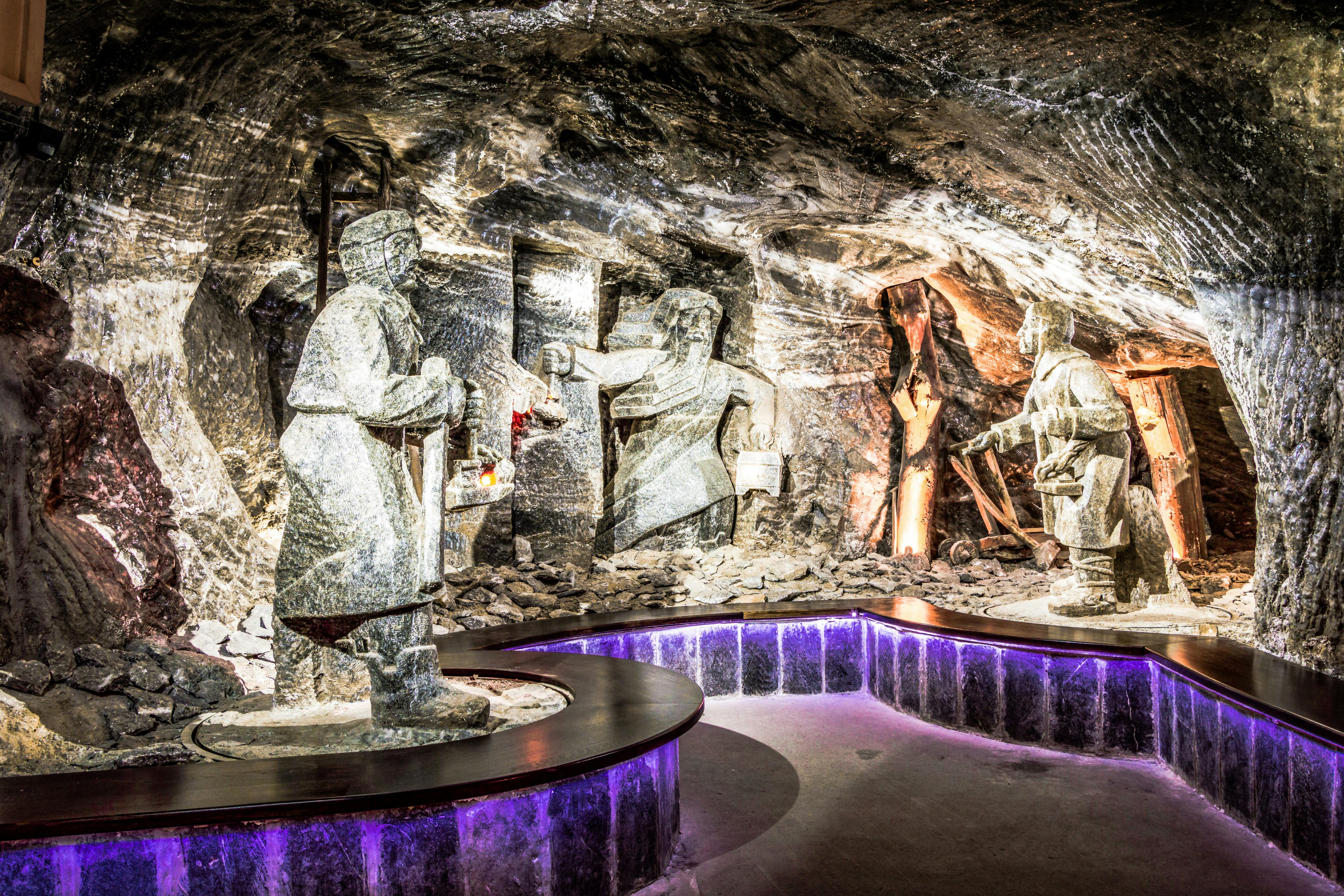 Statues of miners, carved from rock salt, in an underground cave illuminated by purple and white lighting.