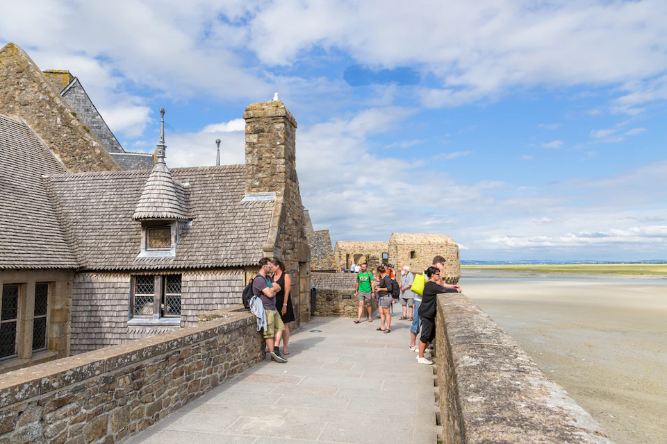 People standing and sitting on a stone pathway overlooking a scenic view near historic stone buildings on a clear, sunny day.