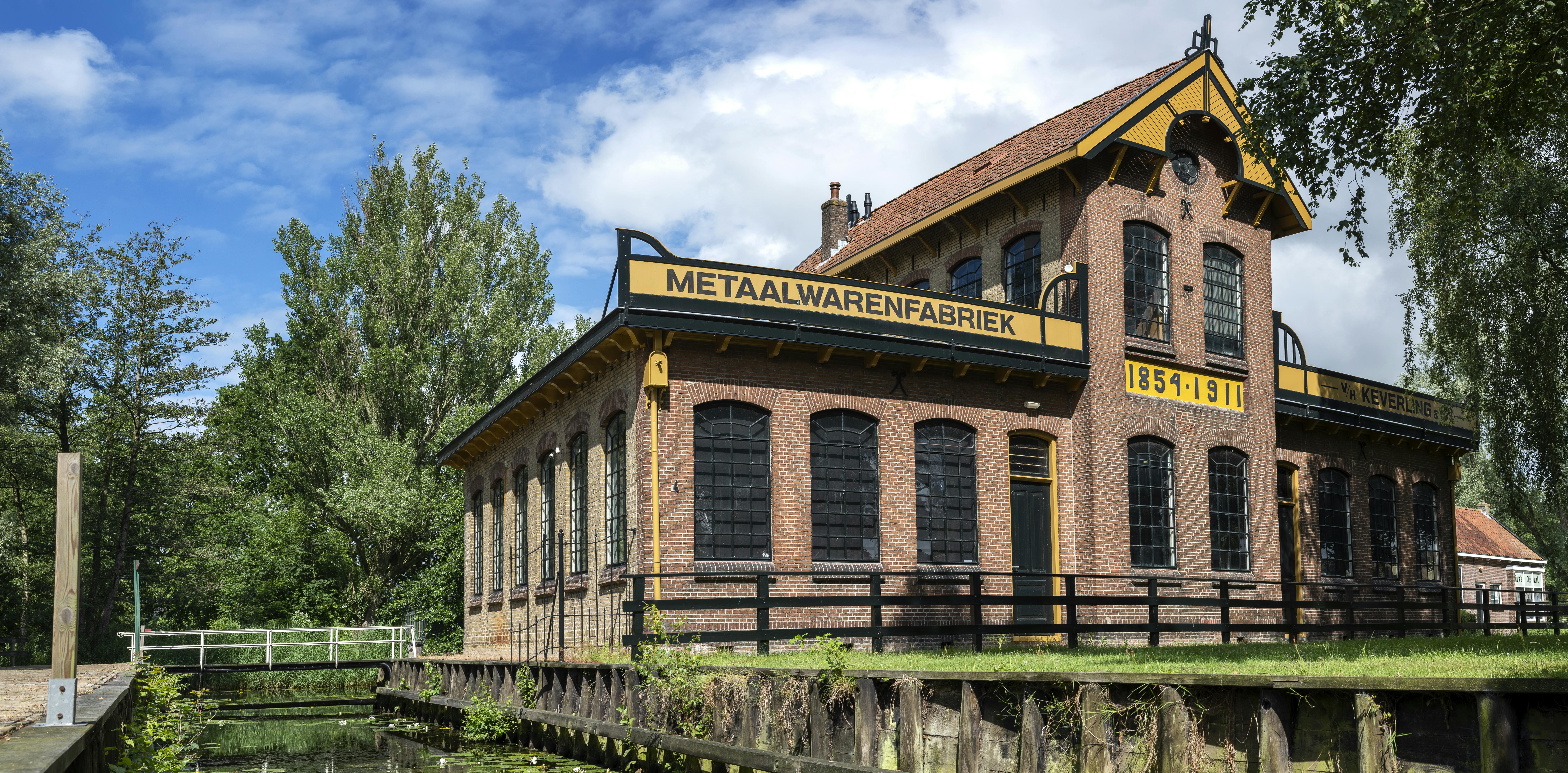 Кирпичное промышленное здание с арочными окнами с надписью «Metaalwarenfabriek» рядом с каналом под частично облачным небом.