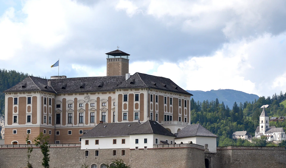 A historic, multi-story building with tall windows and a tower, set against a backdrop of trees and a cloudy sky.