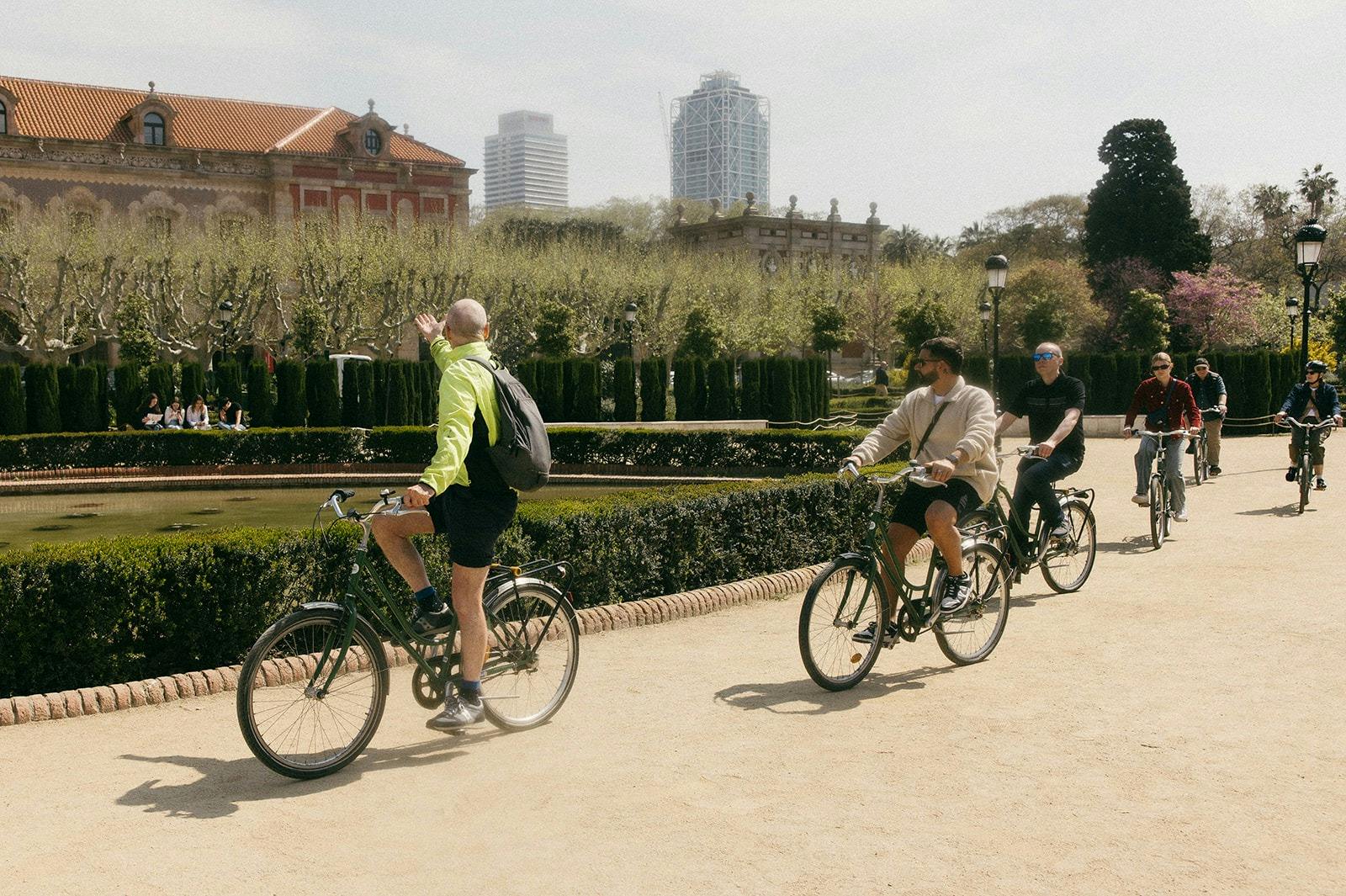 Trois personnes à vélo sur un sentier dans un parc avec des arbres, des buissons et des bâtiments en arrière-plan par une journée ensoleillée.