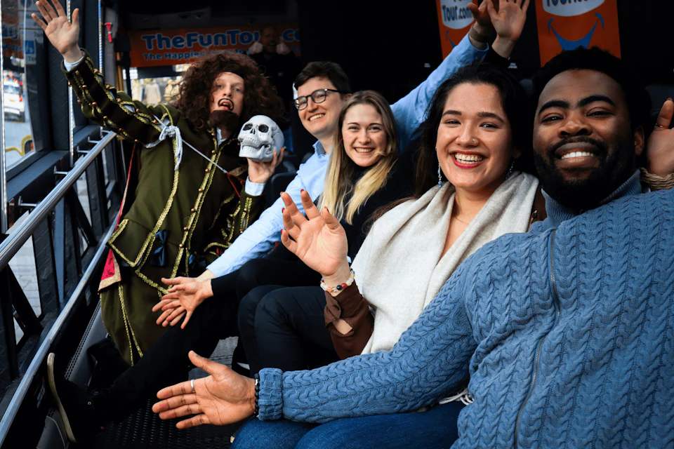 A group of smiling people sitting together, waving at the camera, with a person holding a skull prop on the left.