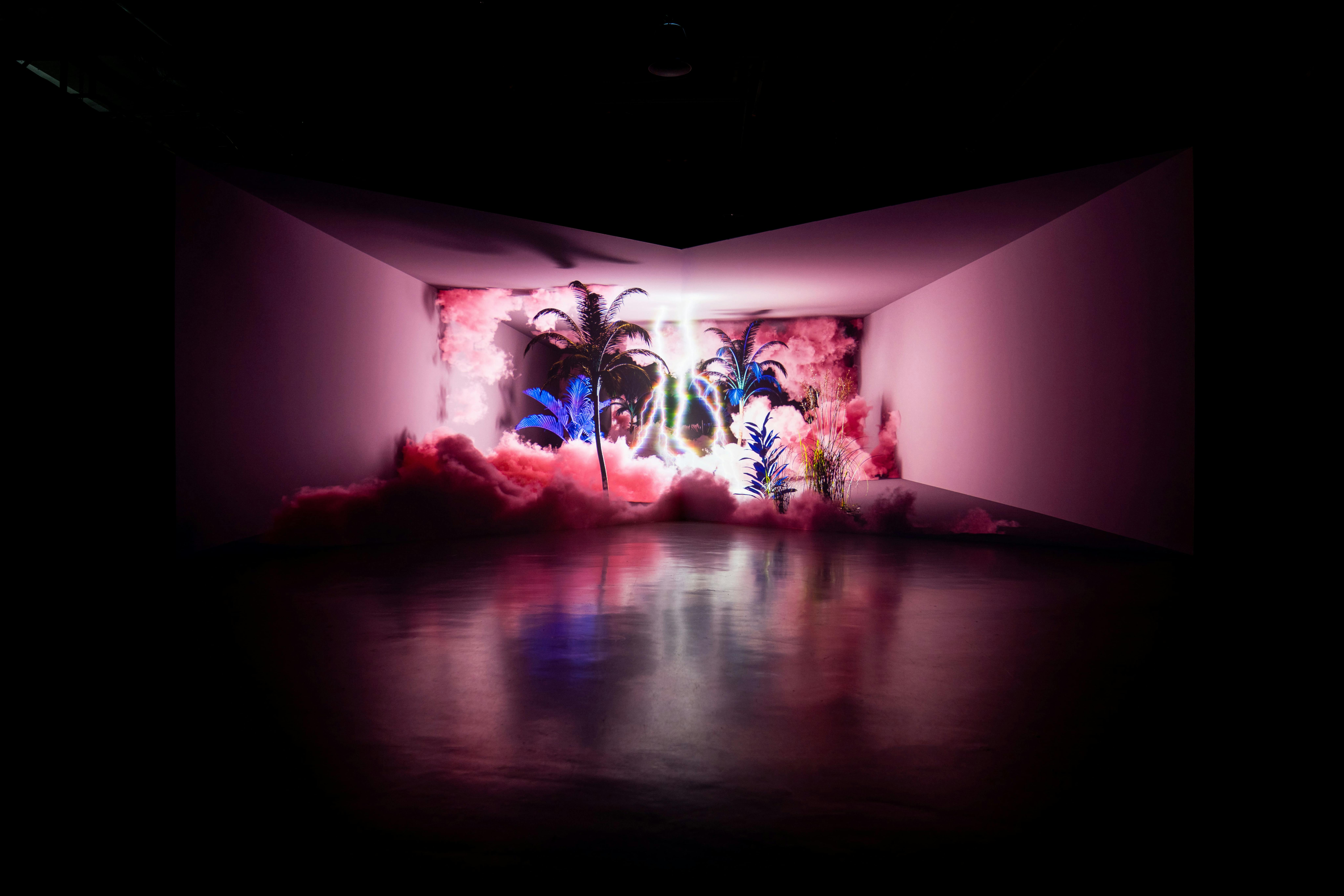 Słabo oświetlony pokój ze świecącymi palmami, różowymi chmurami i odblaskową podłogą. Neony tworzą eteryczną i surrealistyczną atmosferę.