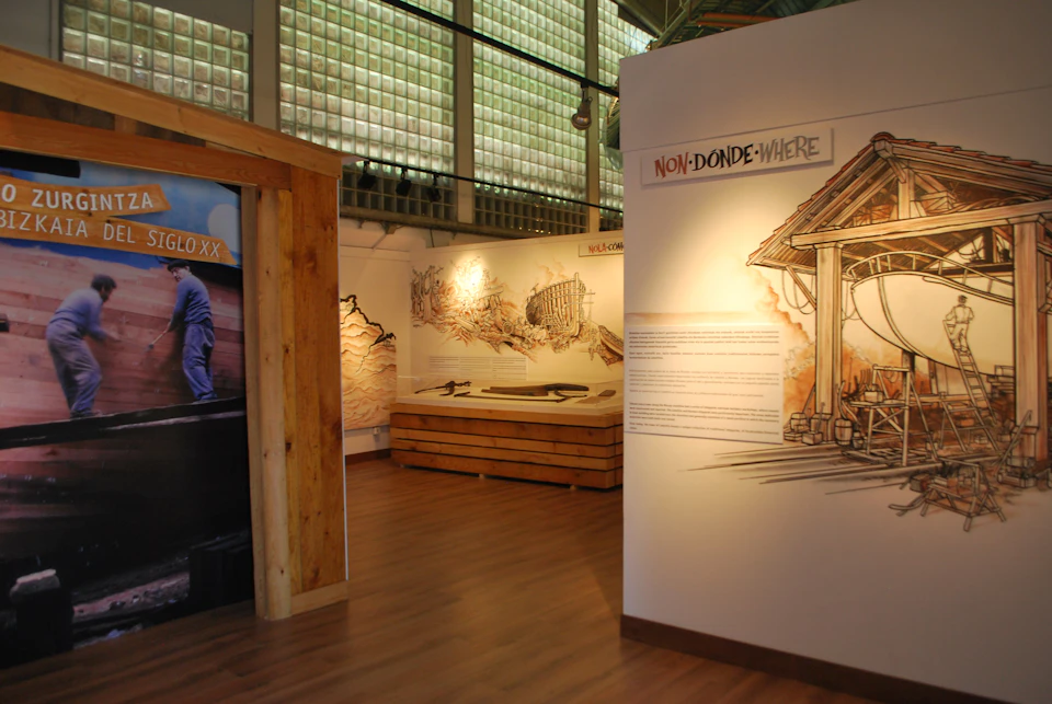 Museum exhibit room featuring illustrations, text panels, and architectural models. Sign reads "NON-DONDE-WHERE" with wooden accents.