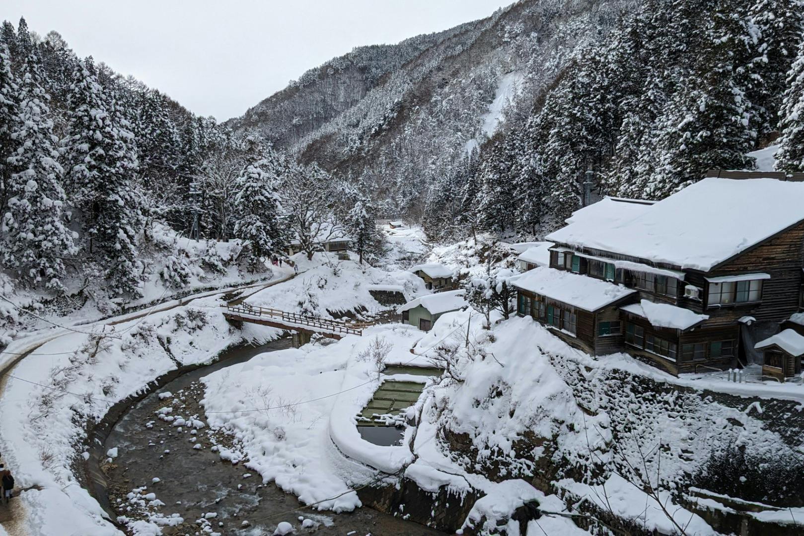 Snow-covered mountain village with a river, wooden bridge, traditional buildings, and surrounding forested hills.