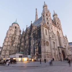 Stor gotisk katedral med höga spiror och invecklade detaljer, omgiven av ett livligt torg med suddiga fotgängare och byggnader.
