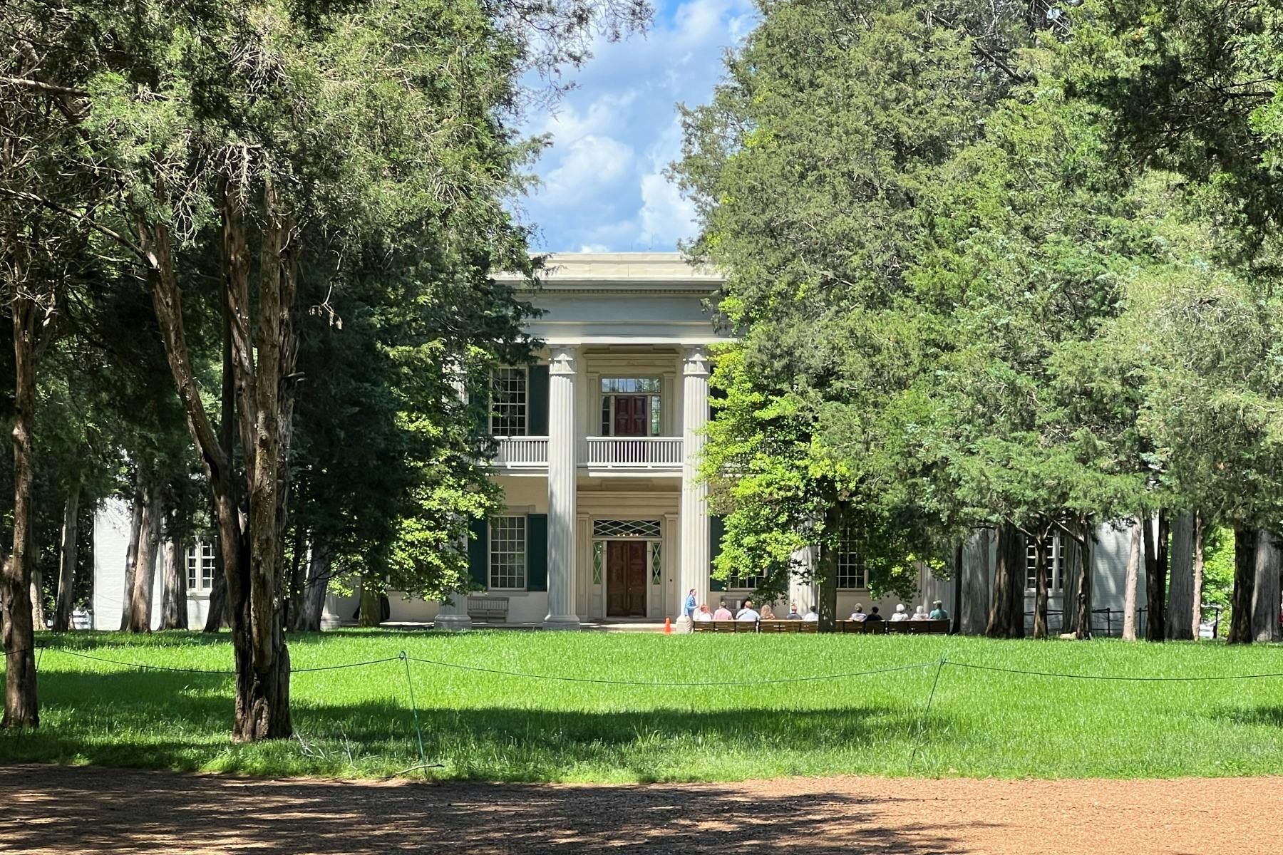 En stor, tvåvånings vit herrgård med pelare, omgiven av höga gröna träd och en välskött gräsmatta under en blå himmel.