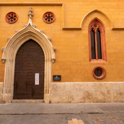 En historisk gul byggnad med välvda trädörrar, röda cirkulära fönster och ett högt smalt fönster. En kakelväggmålning och skylt är synliga.