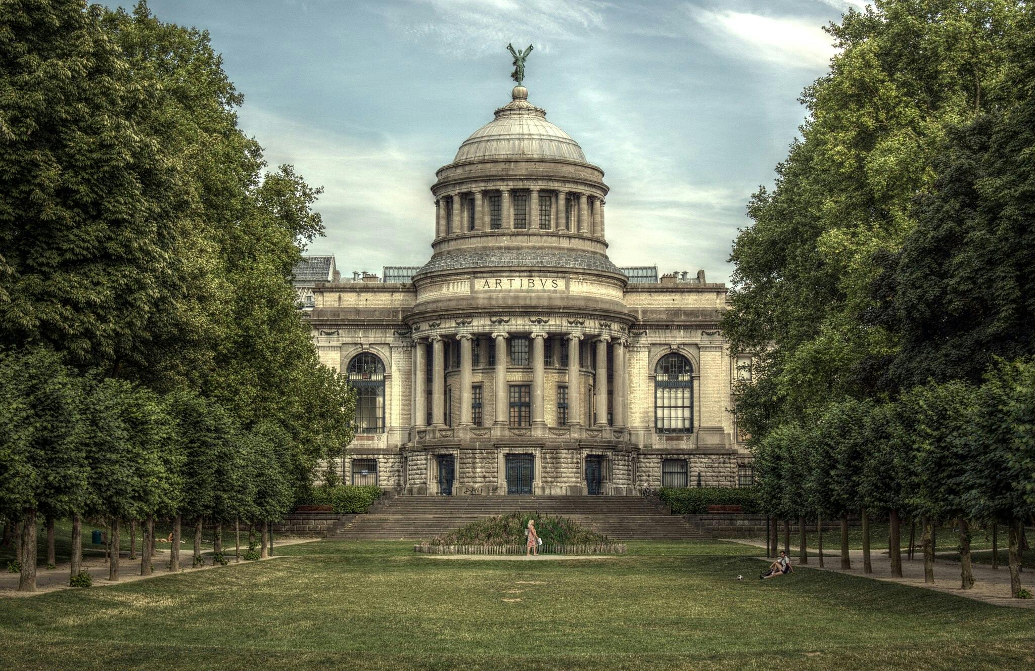 Gran edificio de piedra con columnas y cúpula rodeado de árboles y parque, una persona paseando y otra sentada en la hierba.