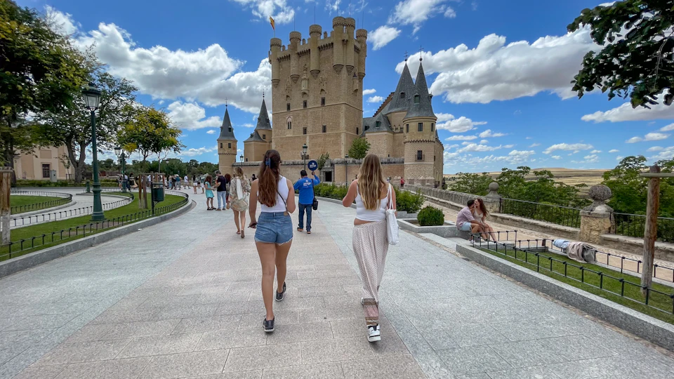 Two women walk towards a historic castle with tall towers under a partly cloudy sky, surrounded by tourists and greenery.