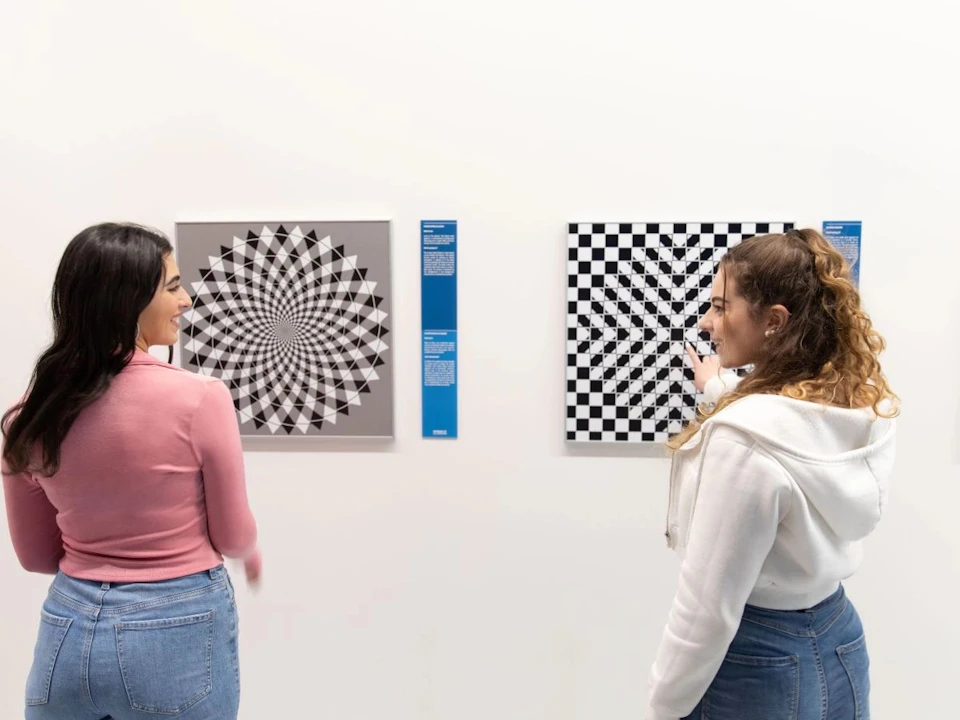 Two women viewing geometric art pieces on a white gallery wall, one with a spiral pattern and the other a checkerboard design.