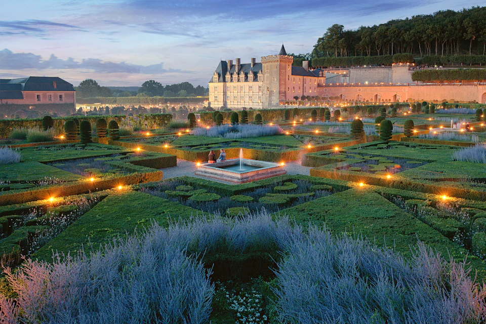 Formal garden with geometric hedges and lit pathways in front of a historic chateau at dusk. Two people sit near a central fountain.