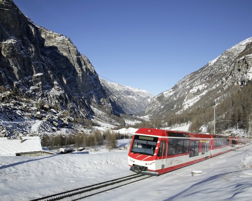 Ένα κόκκινο και άσπρο τρένο ταξιδεύει μέσα σε ένα χιονισμένο ορεινό τοπίο κάτω από έναν καταγάλανο ουρανό.