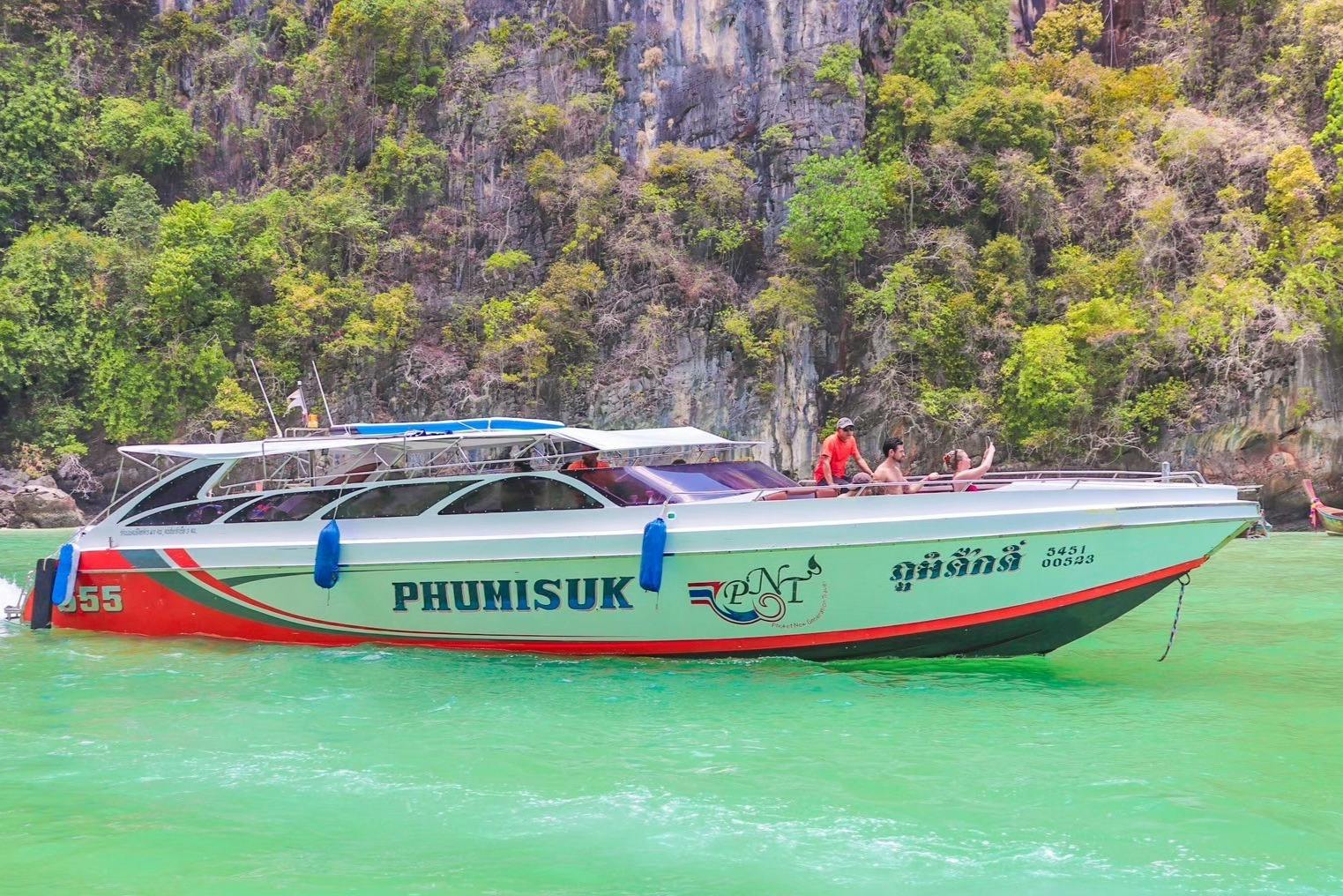 Witte speedboot genaamd &#39;PHUMISUK&#39; met mensen aan boord rondvaart langs een steile, weelderig groene klif over smaragdgroen water.