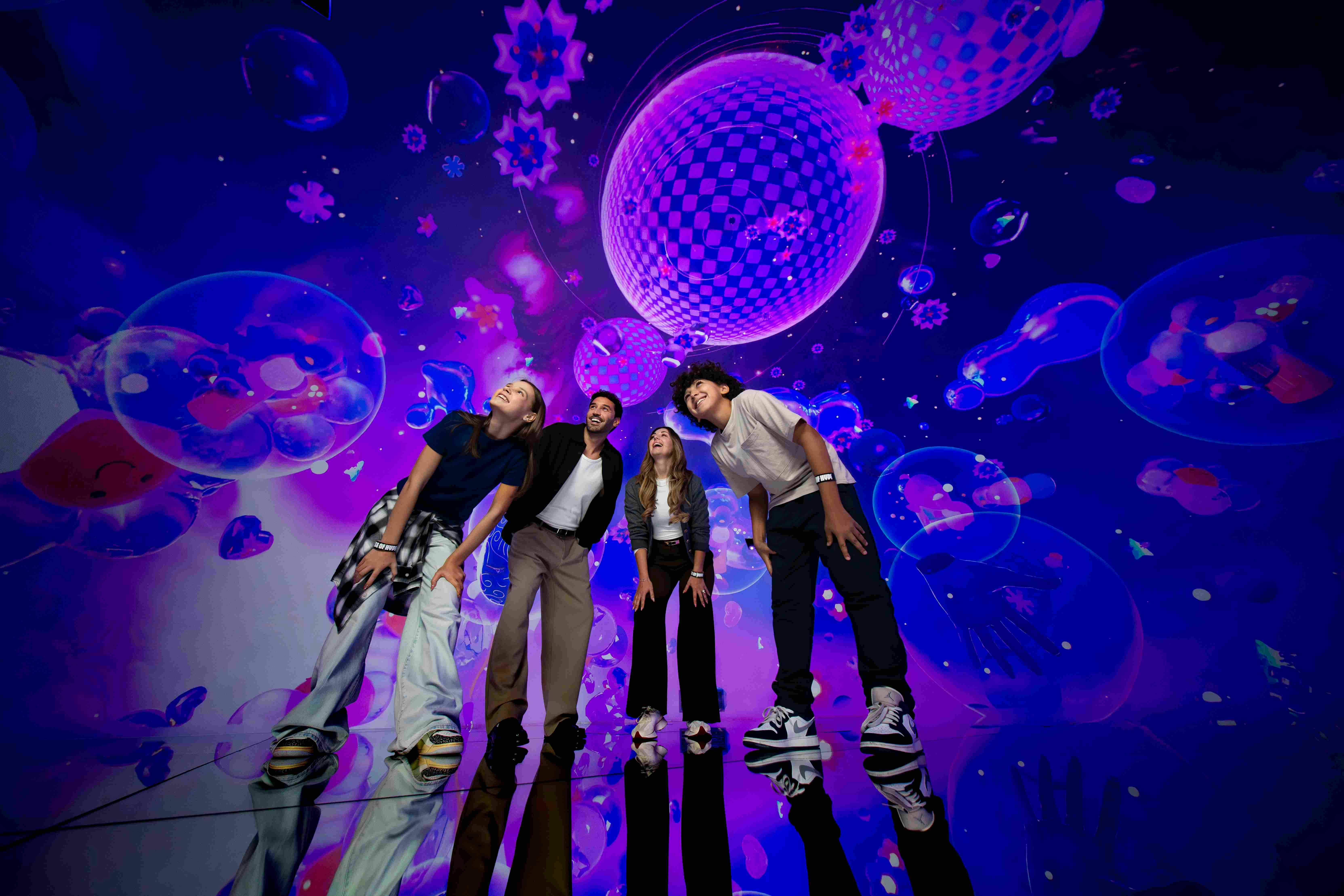 A group of four people stands on a reflective floor under a vivid digital display with purple and pink spherical patterns.