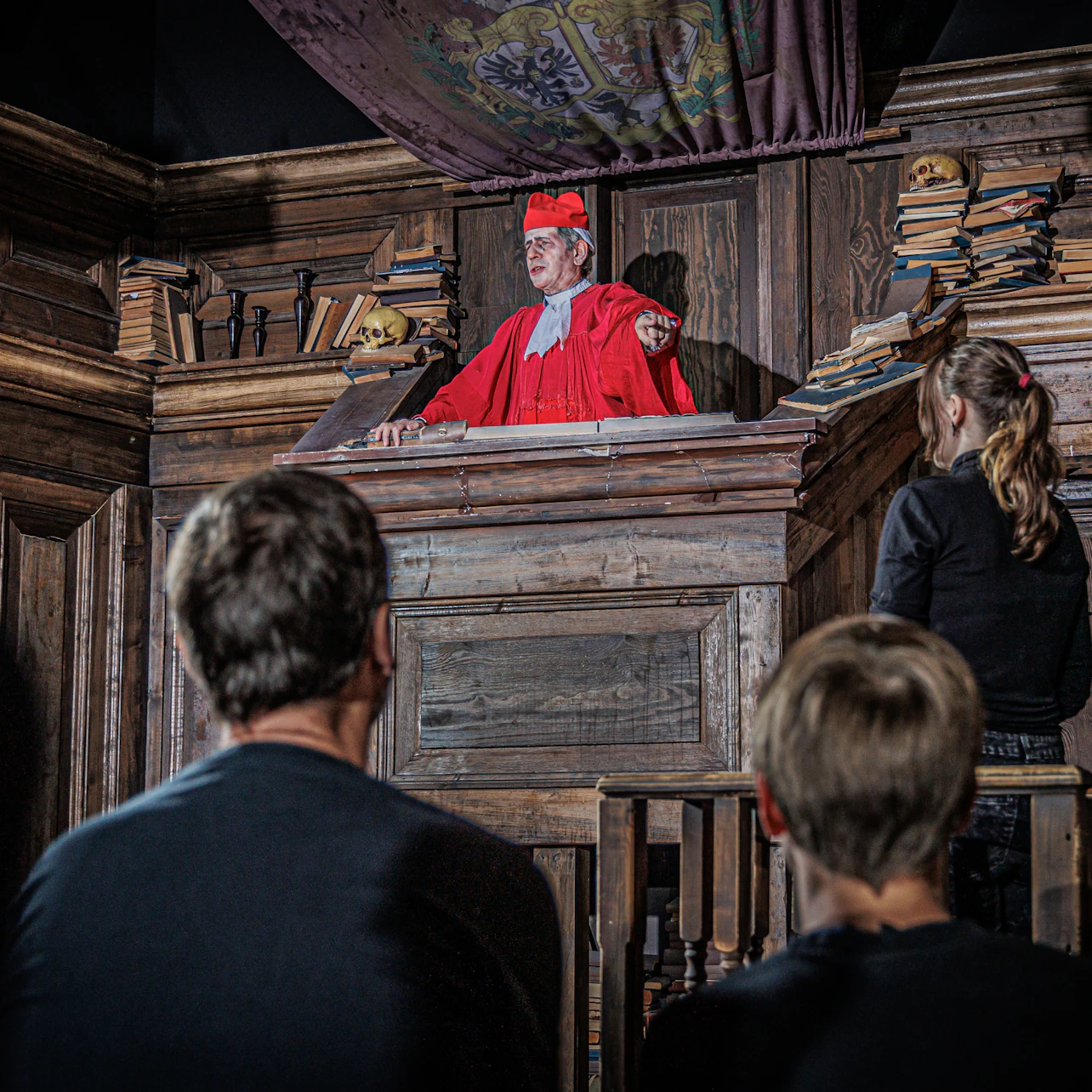 A man in red robes and cap stands in a wooden pulpit, addressing three onlookers. Shelves with books are visible in the background.