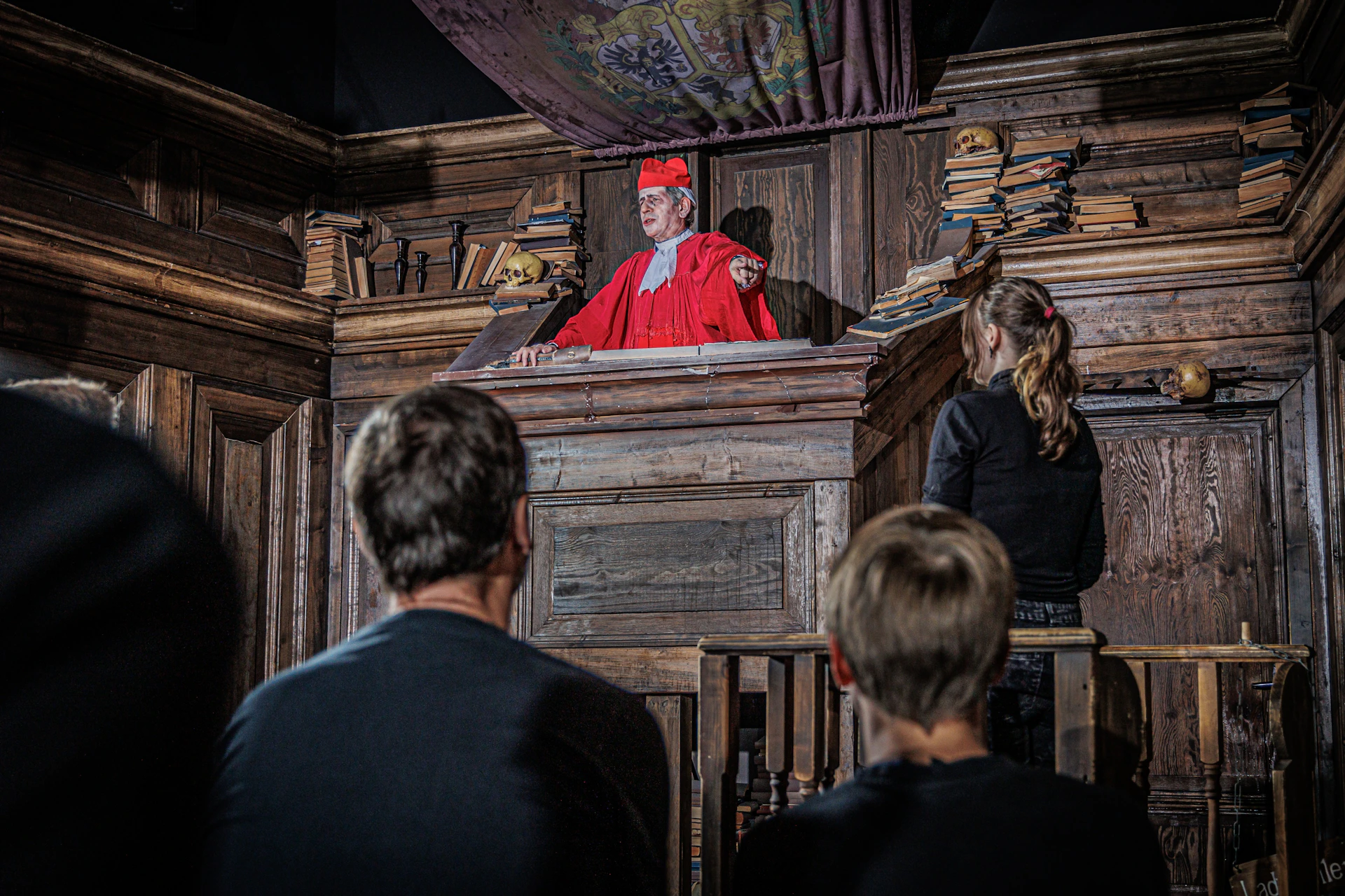 A man in red robes and cap stands in a wooden pulpit, addressing three onlookers. Shelves with books are visible in the background.
