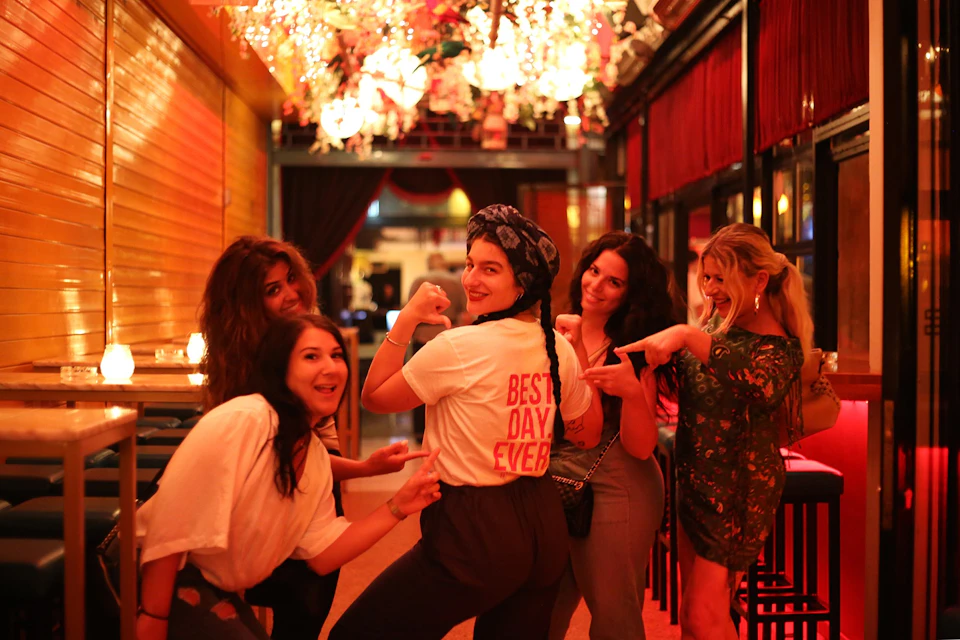 A group of five people in a warmly lit space, with one in a "Best Day Ever" shirt, posing playfully together.