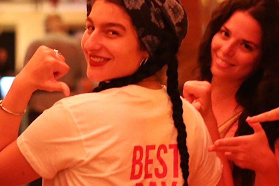 A group of five people in a warmly lit space, with one in a "Best Day Ever" shirt, posing playfully together.