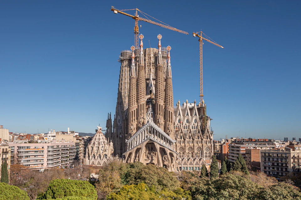 A large, ornate basilica under construction with cranes, surrounded by trees and urban buildings under a clear blue sky.