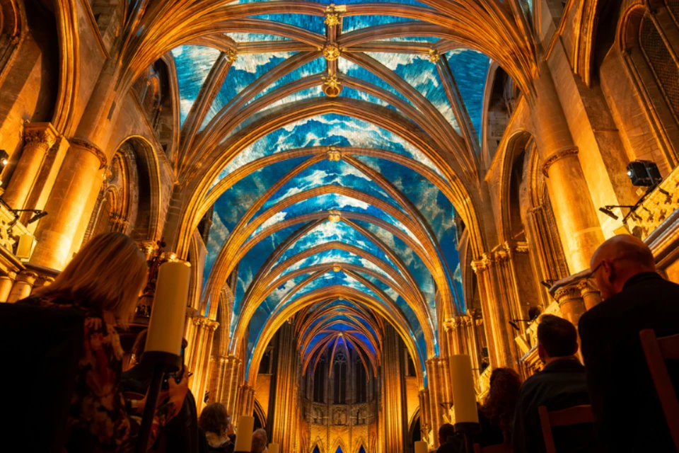 A cathedral interior with illuminated golden arches and a ceiling displaying a vibrant blue sky with clouds.