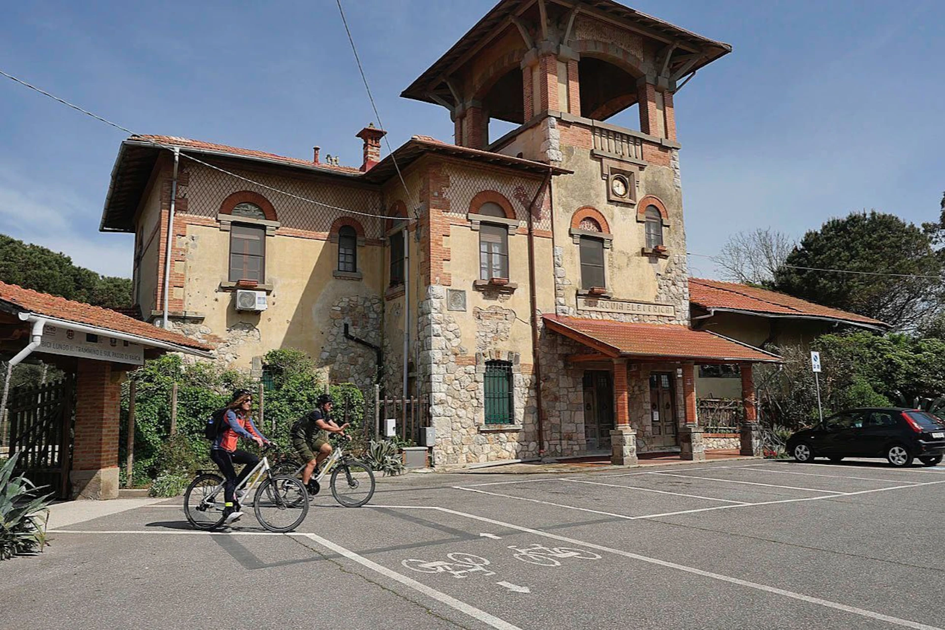 Two people ride bicycles past a rustic, multi-story building with arched windows and a red-tiled roof.