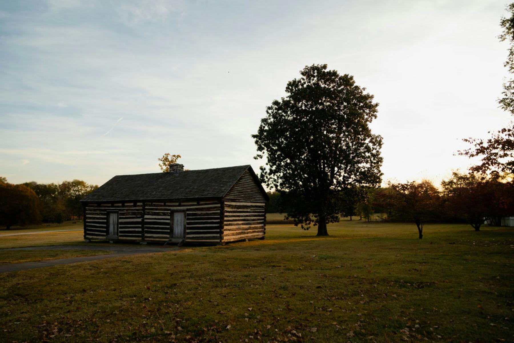 A rustic log cabin stands beside a large tree in a grassy field under a partly cloudy sky at sunset.